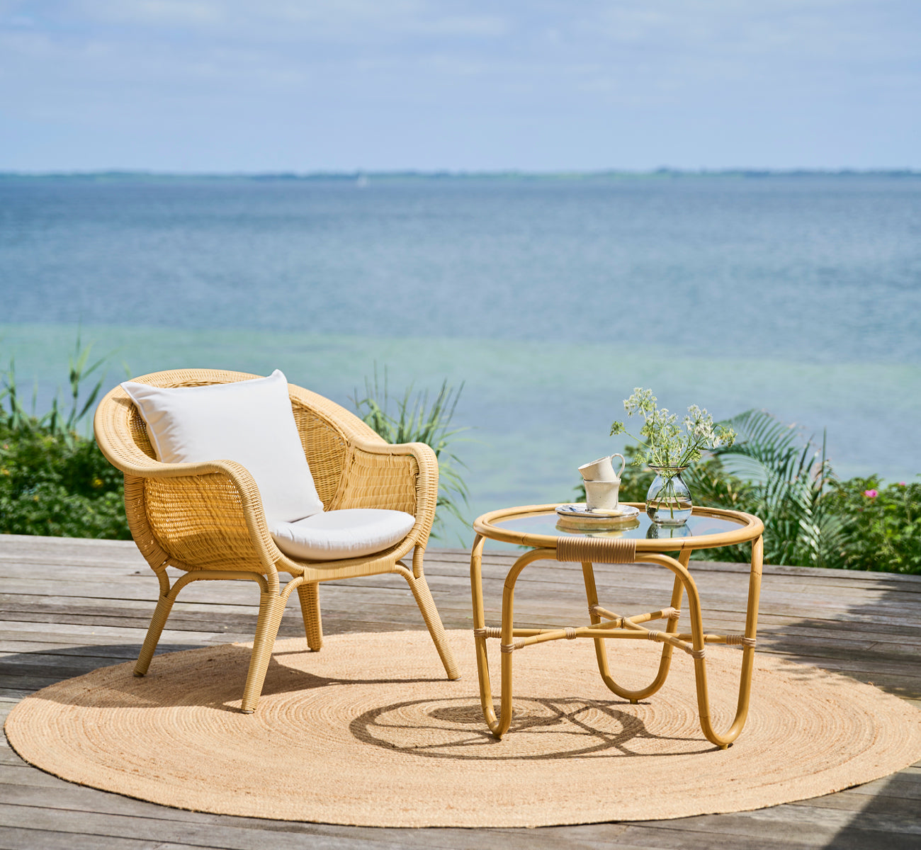 This is a lifestyle image of Madame Lounge Chair in natural core weave ArtFibre Aluminium, on an open coastal terrace at sunset with sea views and.