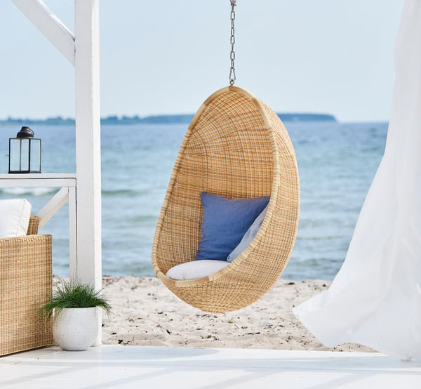 This is a lifestyle image of Hanging Egg Exterior Chair in natural core weave ArtFibre Aluminium, hanging from a white pergola with sandy beach and.