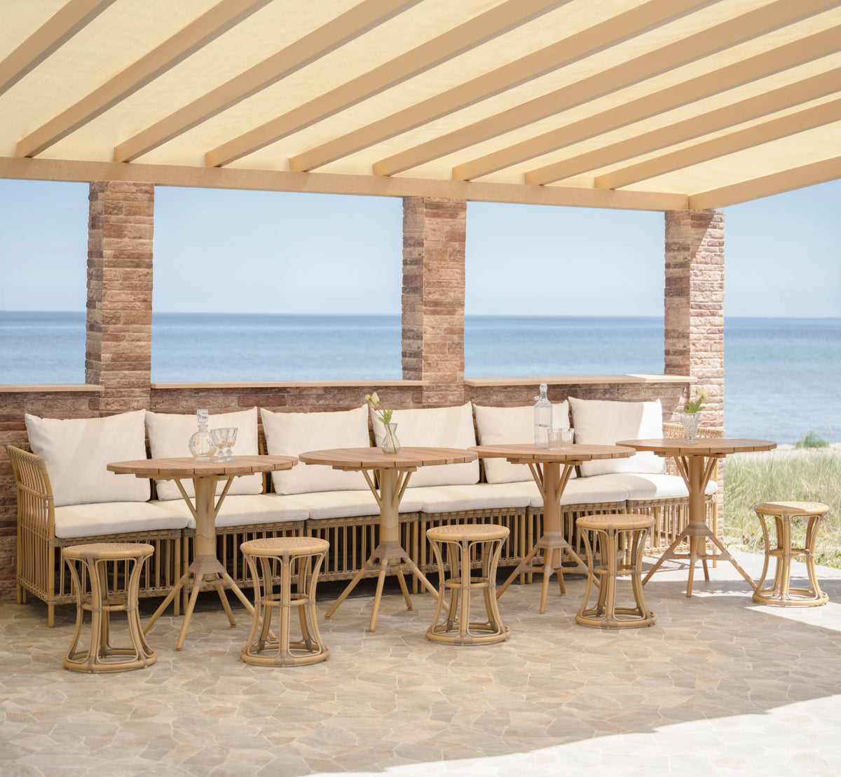 This is a lifestyle image of Bella Exterior Stool in natural aluminium artfibre. Bench with stools and café tables under a pergola.