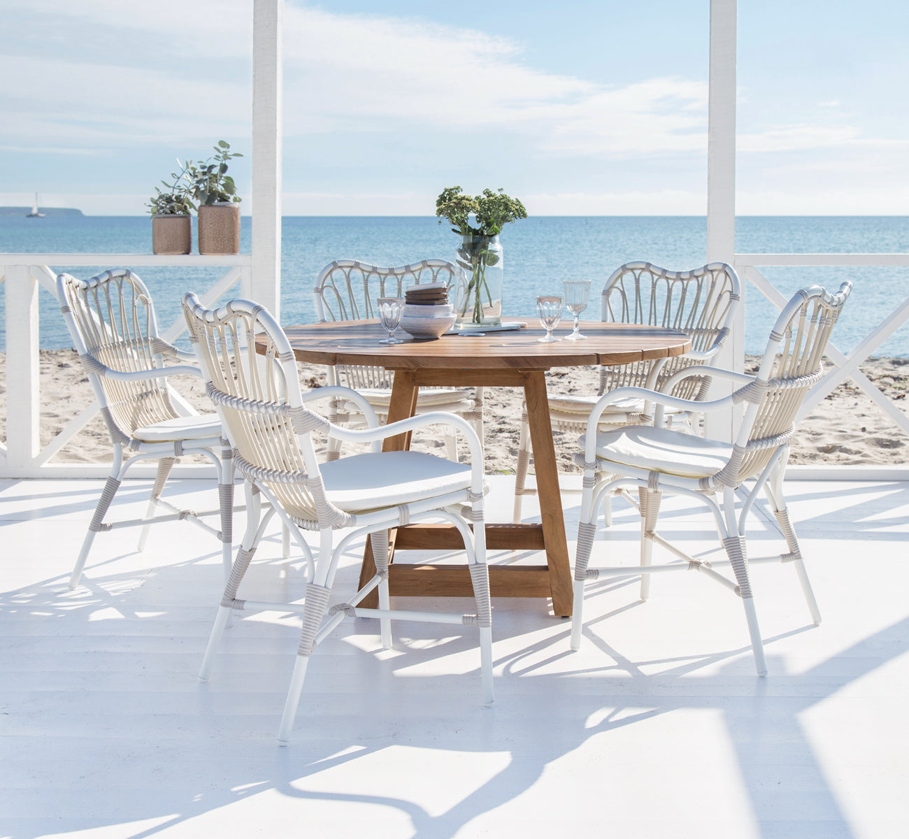 This is a lifestyle image of Margret Exterior Dining Armchair in dove white aluminium artfibre. Beach terrace with teak table and ocean view.