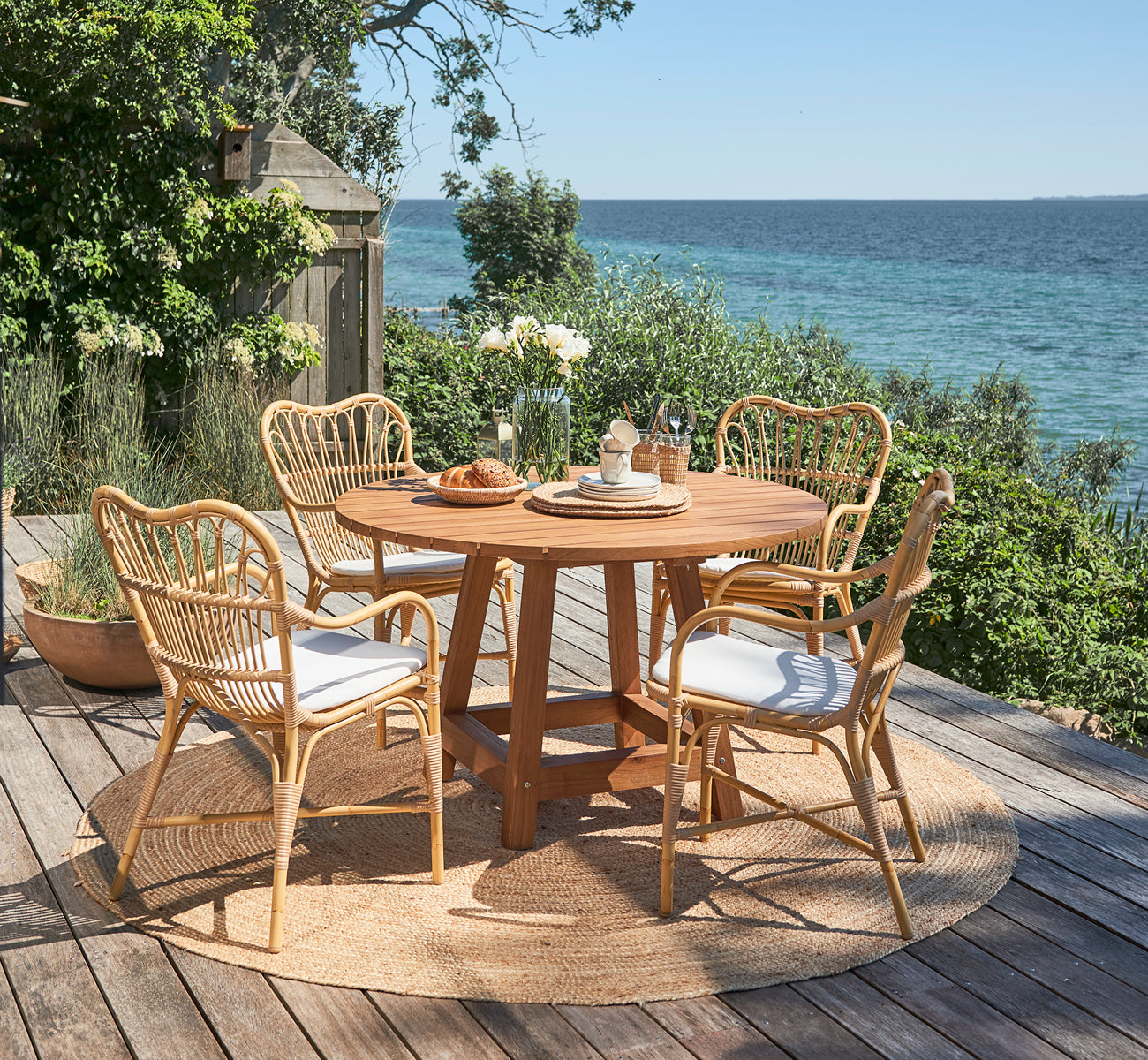 This is a lifestyle image of Margret Exterior Dining Armchair in natural aluminium artfibre. Seaside deck with round teak table and garden plants.