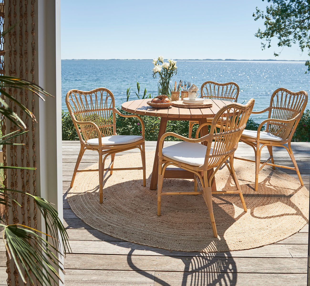 This is a lifestyle image of Margret Exterior Dining Armchair in natural aluminium artfibre. Seaside deck with round teak table and ocean view.