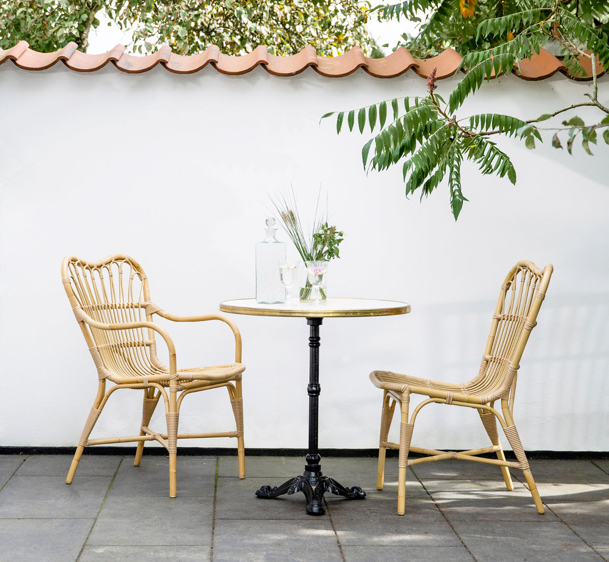 This is a lifestyle image of Margret Exterior Dining Armchair in natural aluminium artfibre. Courtyard with café table under tiled roof overhang.