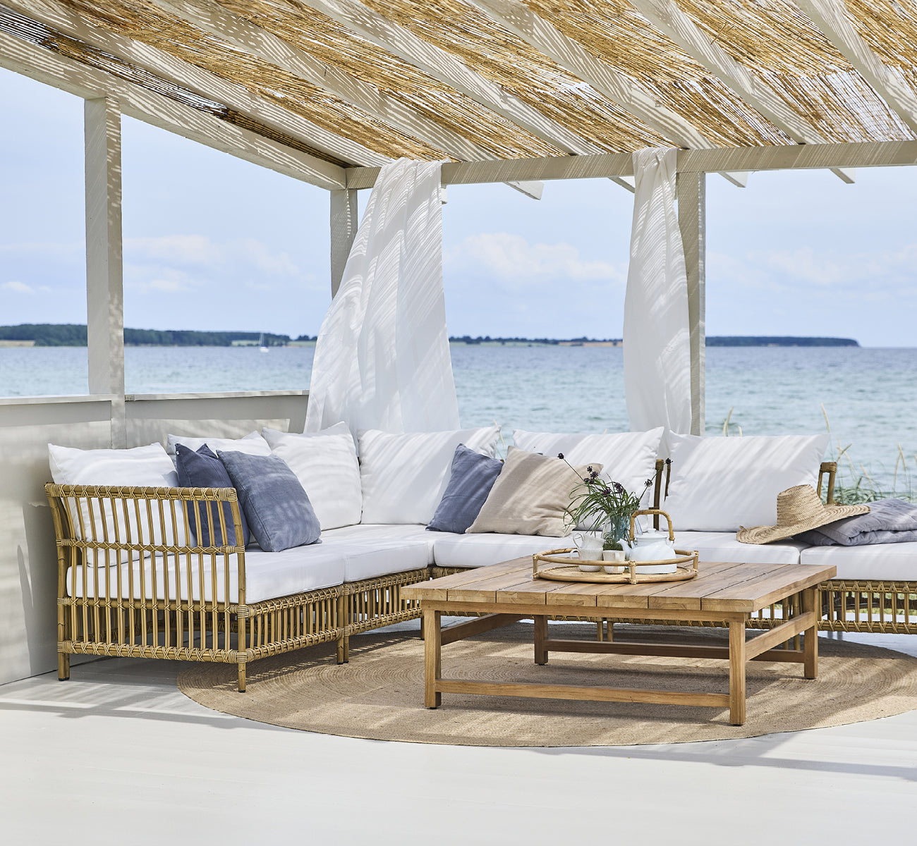 This is a lifestyle image of Maggie Sofa Corner Module in antique aluminium artfibre. Beach pavilion with sofa set and ocean view.