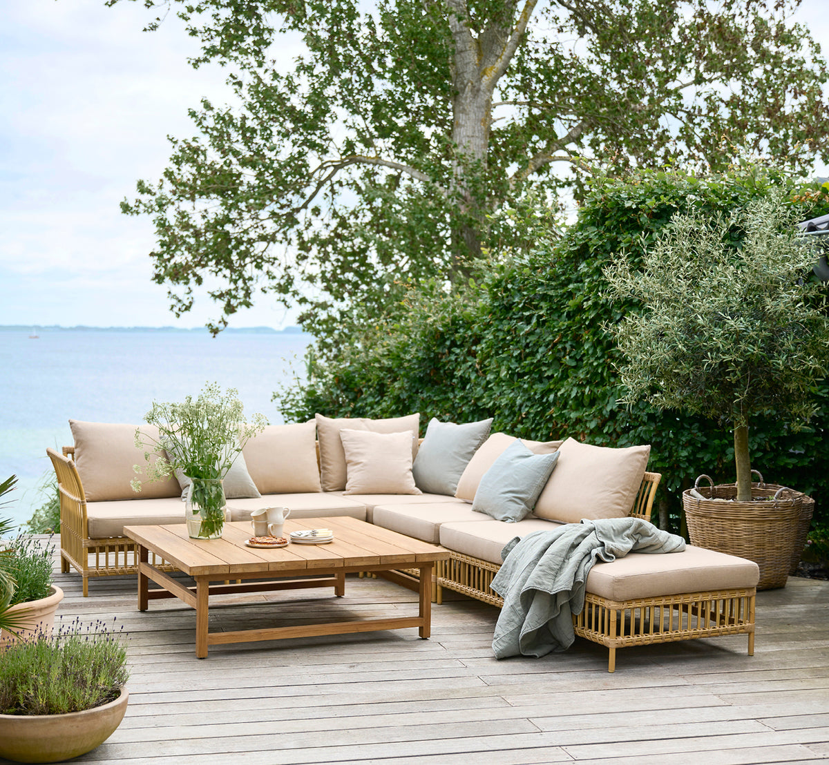 This is a lifestyle image of Maggie Sofa Corner Module in natural aluminium artfibre. Seaside wooden deck with cushions and ocean view.