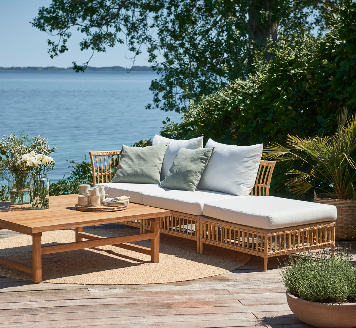 This is a lifestyle image of Maggie Sofa Center Module in natural aluminium artfibre. Seaside deck with cushions, plants, and ocean view.