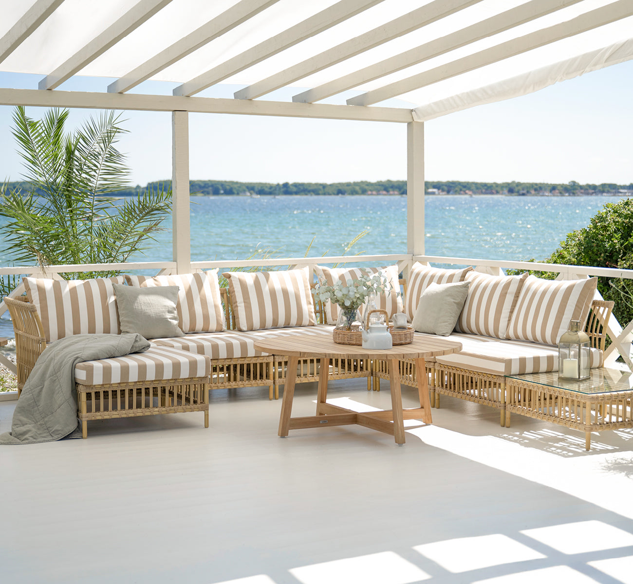 This is a lifestyle image of Maggie Sofa Center Module in natural aluminium artfibre. Covered terrace with striped cushions and sea backdrop.