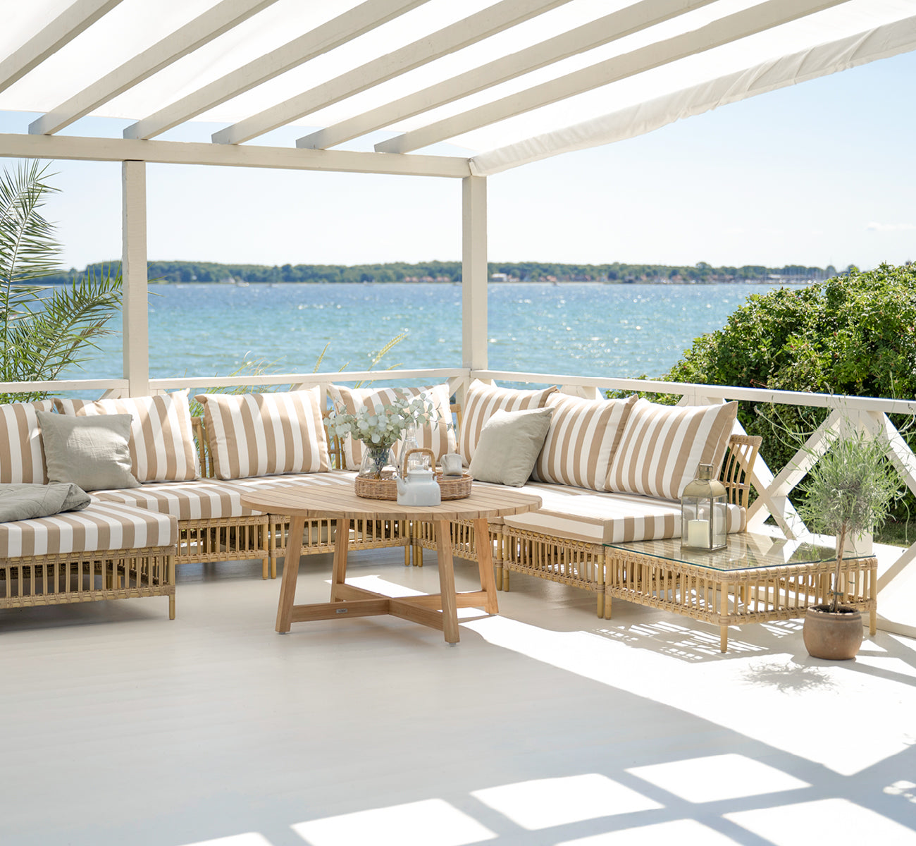 This is a lifestyle image of Maggie Sofa Pouf Module w. Glass in natural aluminium artfibre. Covered terrace with striped cushions and sea view.