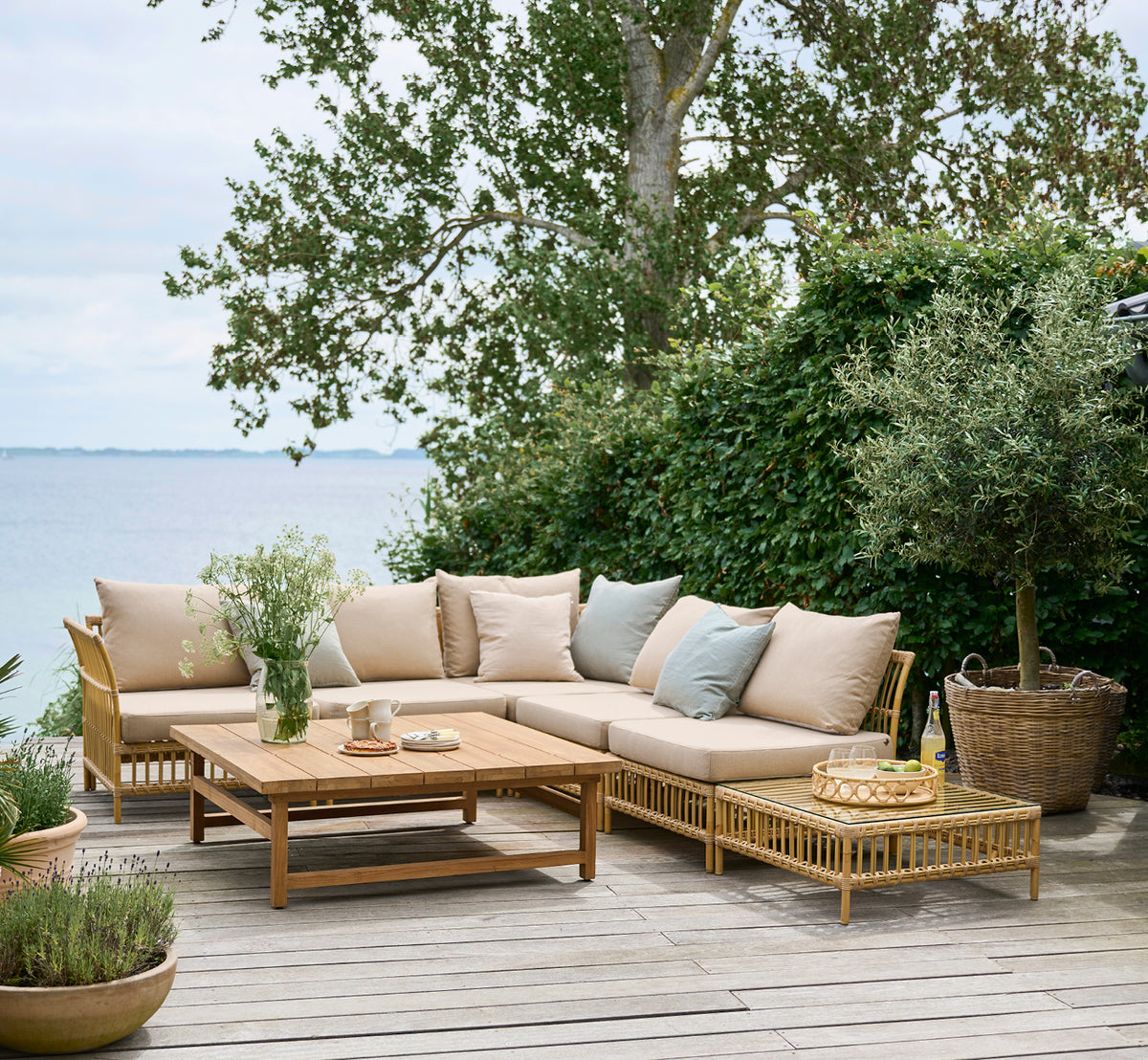 This is a lifestyle image of Maggie Sofa Pouf Module w. Glass in natural aluminium artfibre. Seaside deck with sofa set and greenery around.