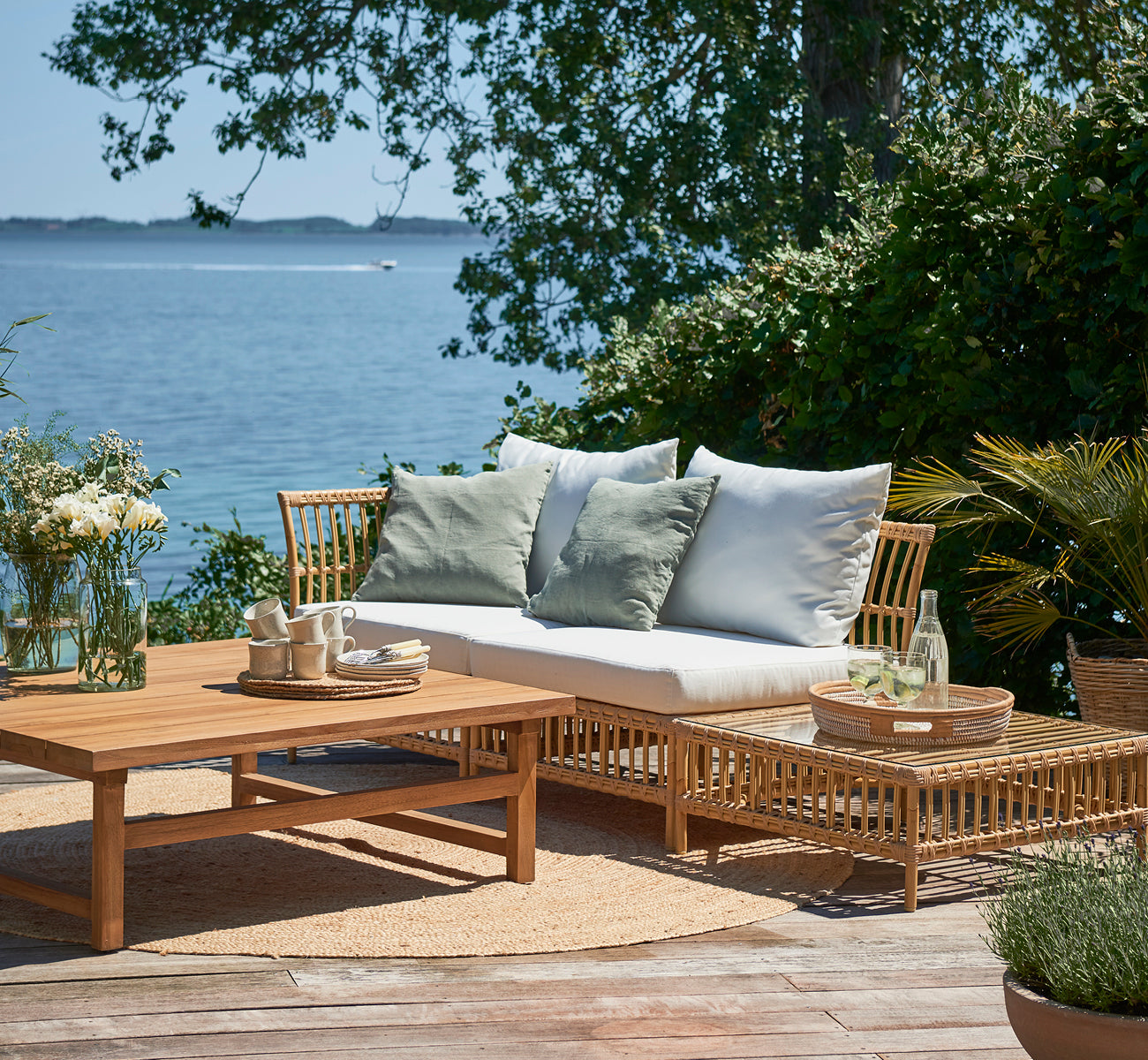 This is a lifestyle image of Maggie Sofa Pouf Module w. Glass in natural aluminium artfibre. Seaside deck with coffee table and green cushions.