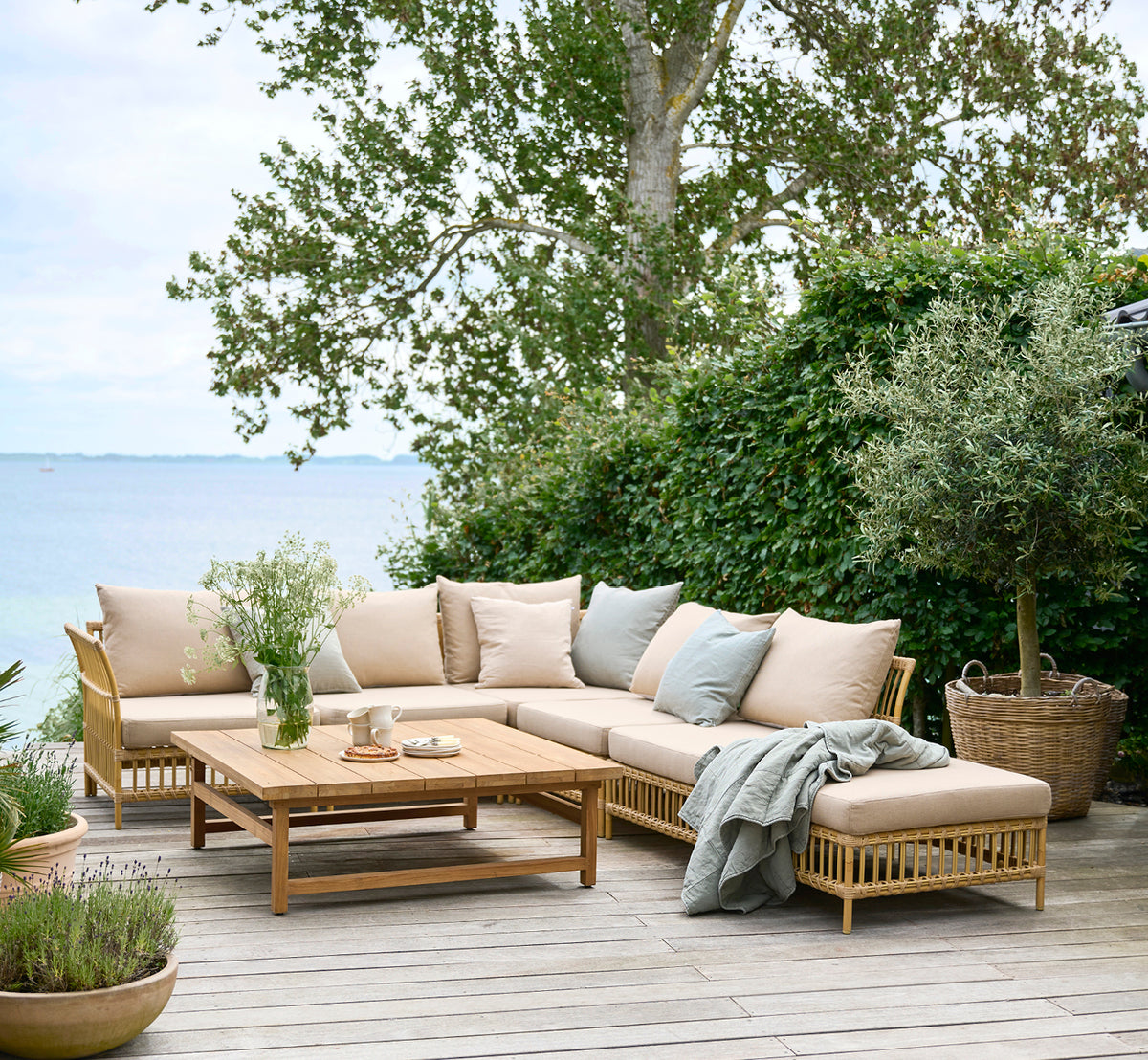 This is a lifestyle image of Maggie Sofa Pouf Module w. Glass in natural aluminium artfibre. Seaside deck with modular sofa and blanket draped.