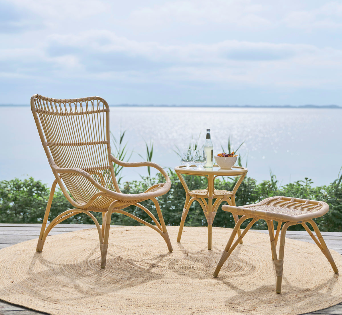 This is a lifestyle image of Monet Exterior Lounge Chair in natural aluminium artfibre. With footstool on a deck overlooking the sea.