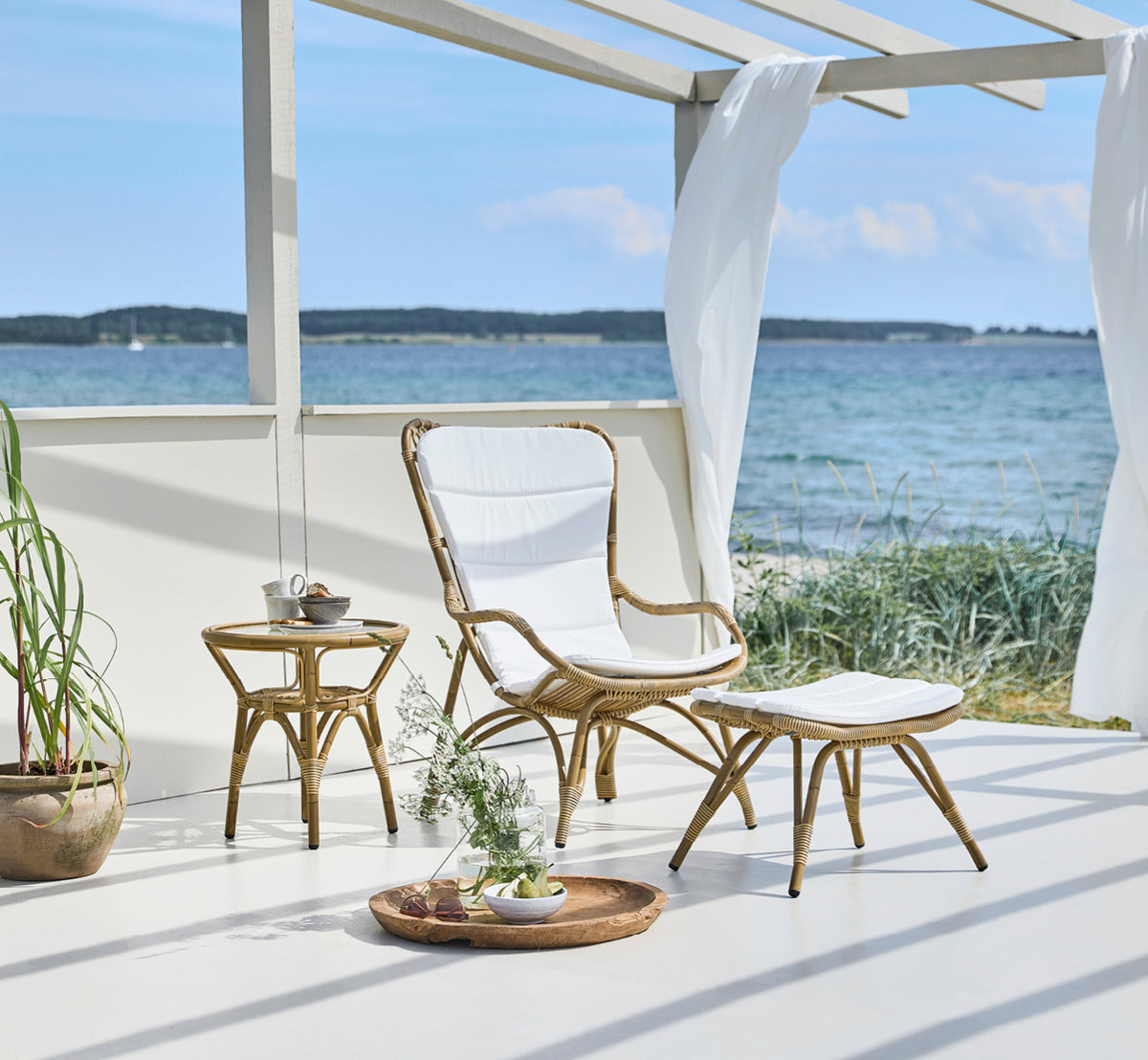 This is a lifestyle image of Monet Exterior Footstool in antique aluminium artfibre. On a seaside pavilion with cushioned chair.