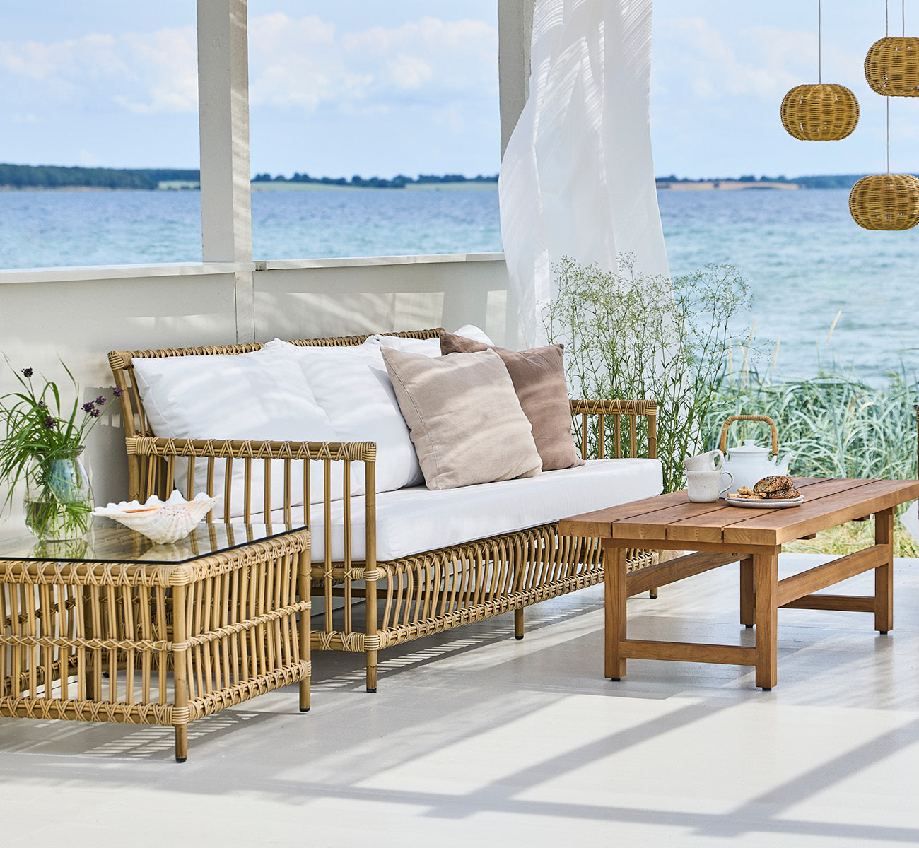 This is a lifestyle image of Caroline Exterior Sofa in antique Aluminium ArtFibre, on a waterside wooden deck with coastal greenery and sea views.