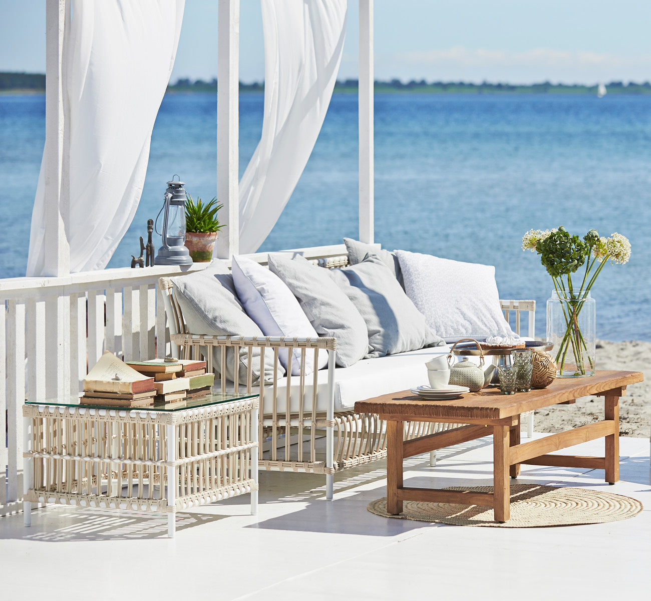 This is a lifestyle image of Caroline Exterior Sofa in dove White Aluminium ArtFibre, on a waterside wooden deck with coastal greenery and sea views.