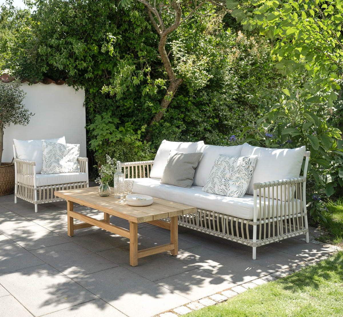 This is a lifestyle image of Caroline Exterior Sofa in dove White Aluminium ArtFibre, on a waterside wooden deck with coastal greenery and sea views.