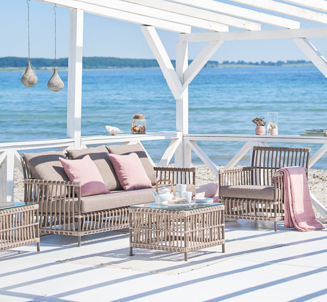 This is a lifestyle image of Caroline Exterior Sofa in moccaccino Aluminium ArtFibre, on a waterside wooden deck with coastal greenery and sea views.