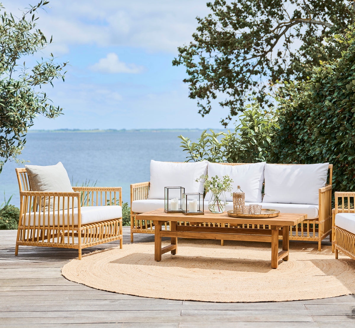 This is a lifestyle image of Caroline Exterior Sofa in natural Aluminium ArtFibre, on a waterside wooden deck with coastal greenery and sea views.