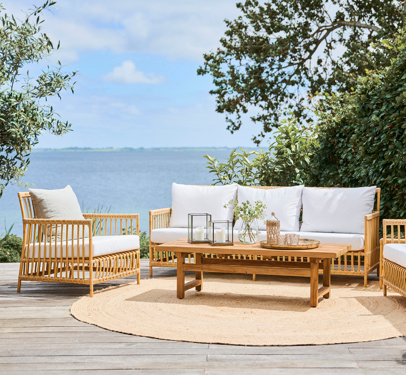 This is a lifestyle image of Caroline Exterior Sofa in natural Aluminium ArtFibre, on a waterside wooden deck with coastal greenery and sea views.