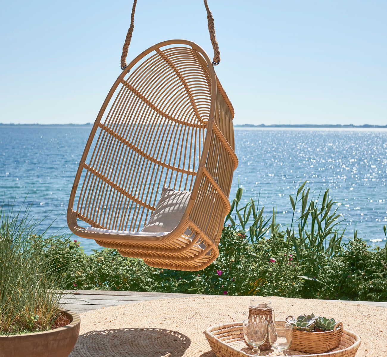 This is a lifestyle image of Renoir Exterior Hanging Chair in natural aluminium artfibre. Hanging on a seaside terrace with tray and succulents below.