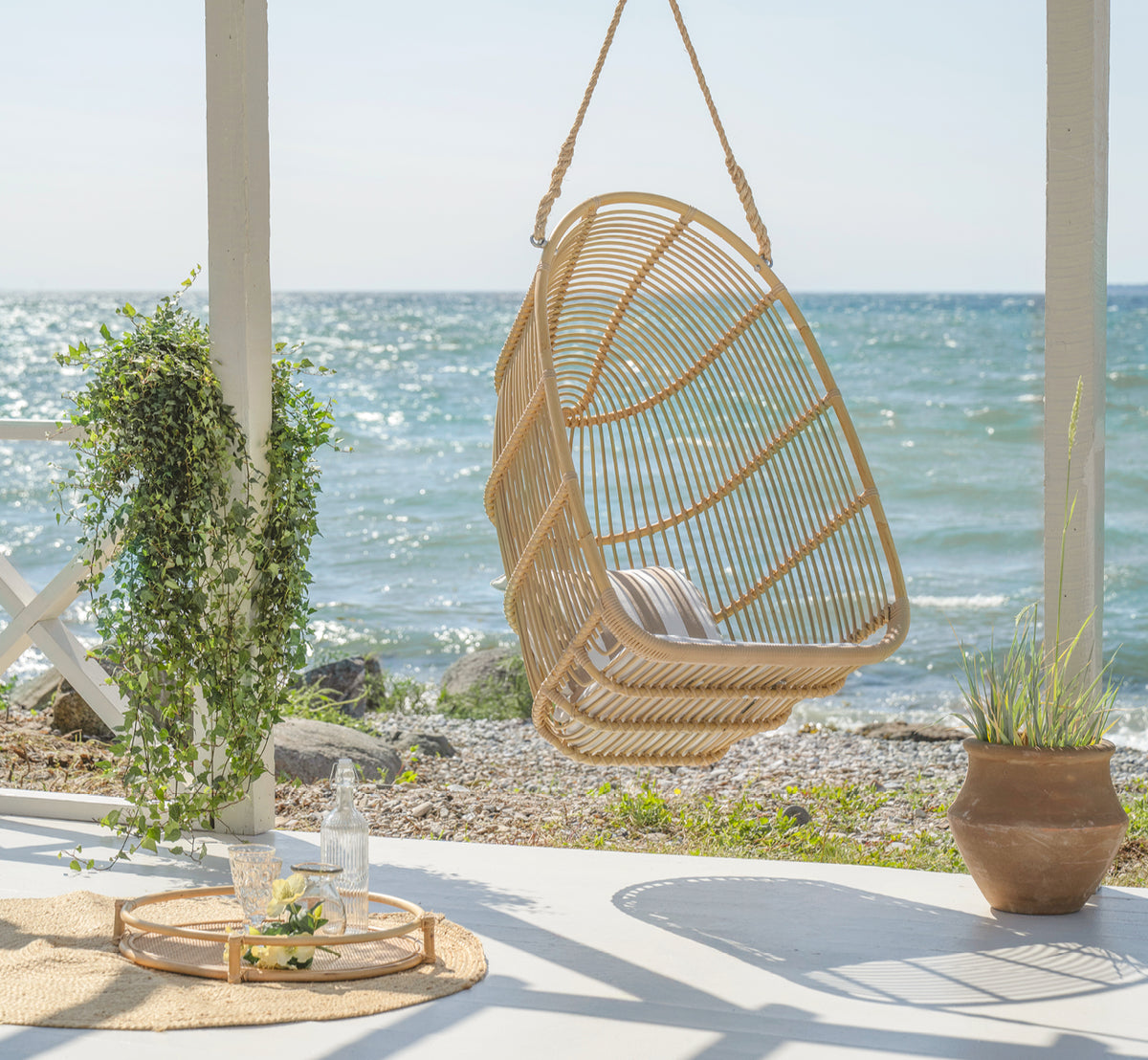 This is a lifestyle image of Renoir Exterior Hanging Chair in natural aluminium artfibre. Hanging on a seaside terrace with tray and succulents below.