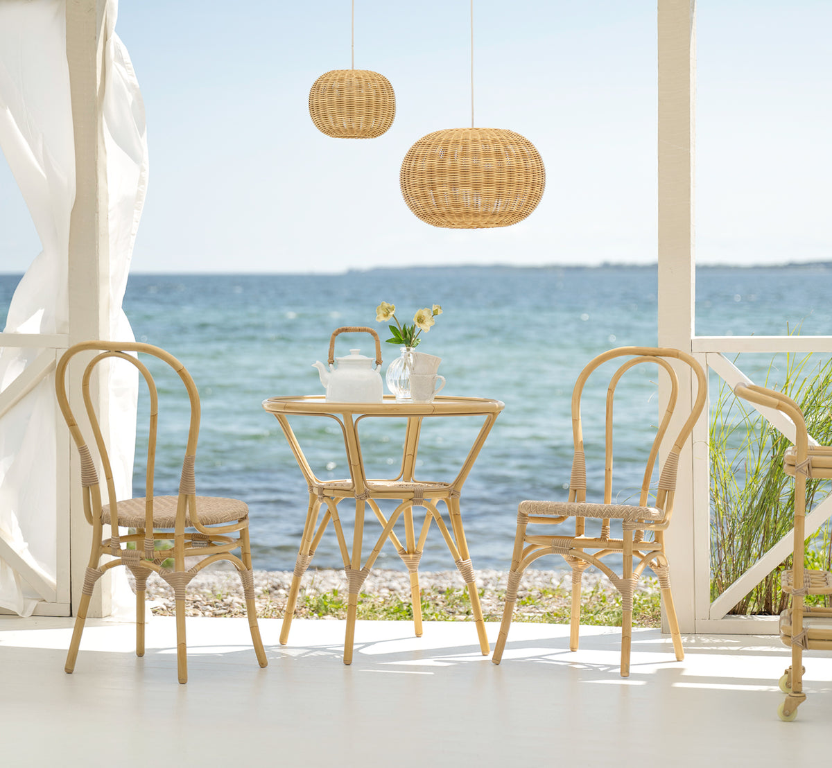 This is a lifestyle image of Tony Exterior Café Table in natural aluminium artfibre. On a seaside terrace with chairs, pendant lamp, and ocean view.