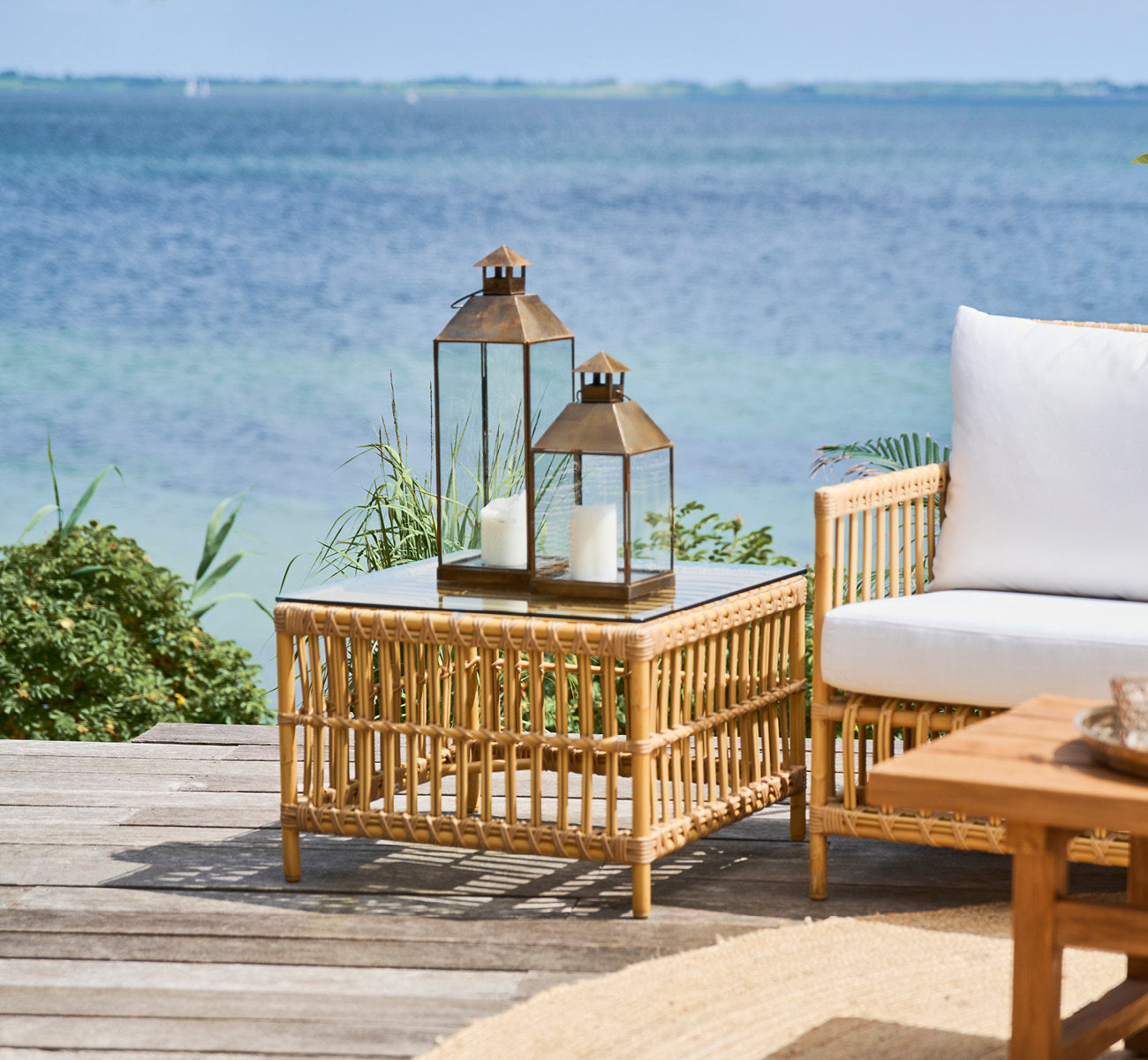 This is a lifestyle image of Caroline Exterior Table in natural Aluminium ArtFibre, on a waterside wooden deck with coastal greenery and sea views.