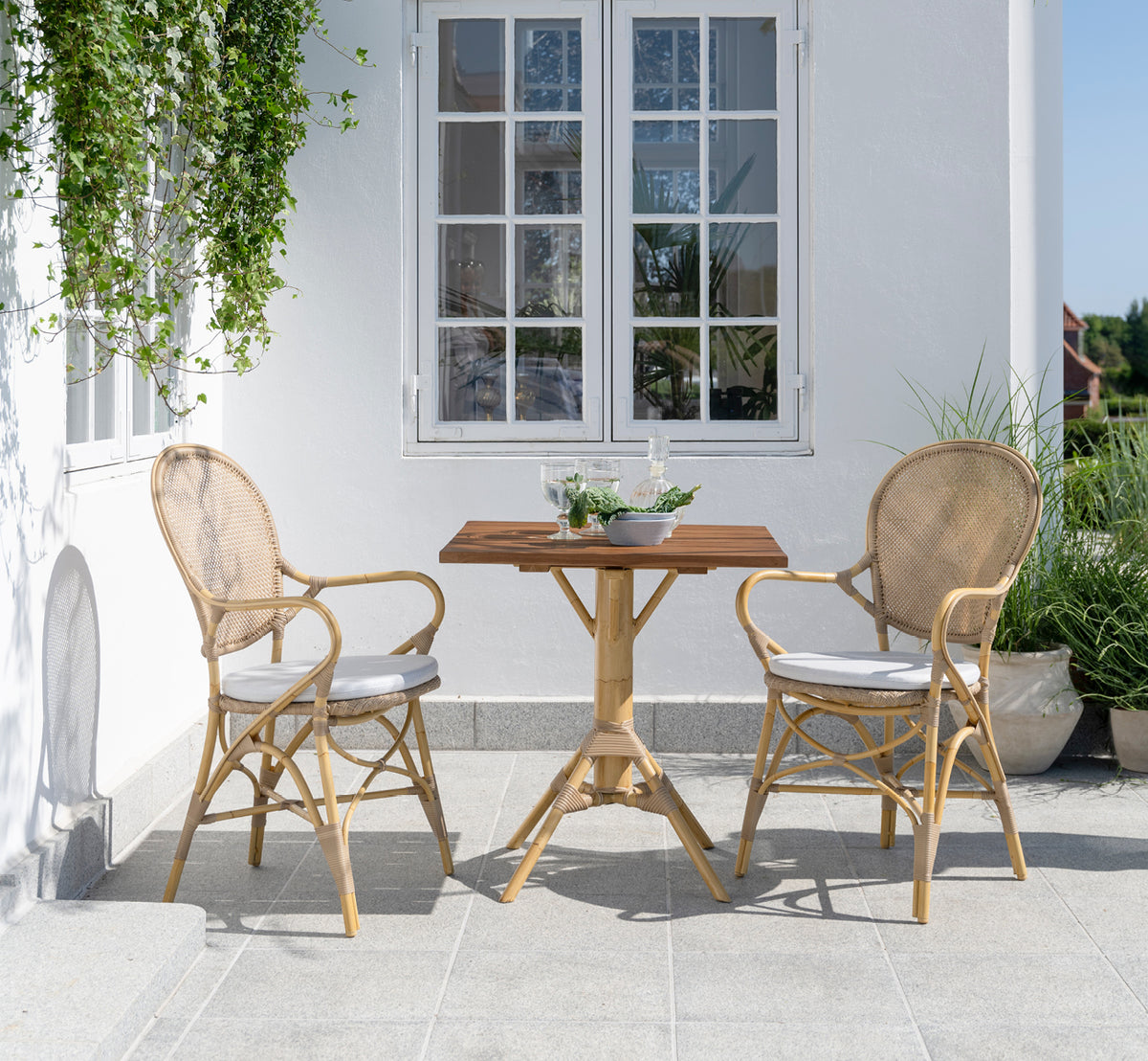 This is a lifestyle image of Nicole Cafe Table in natural aluminium artfibre. On a stone patio with Ofelia chairs and greenery.