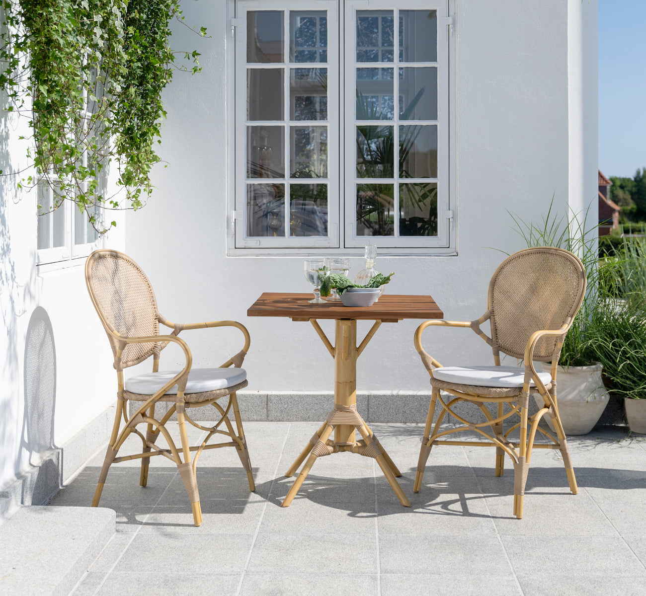 This is a lifestyle image of Nicole Cafe Table in natural aluminium artfibre. On a stone patio with Ofelia chairs and greenery.