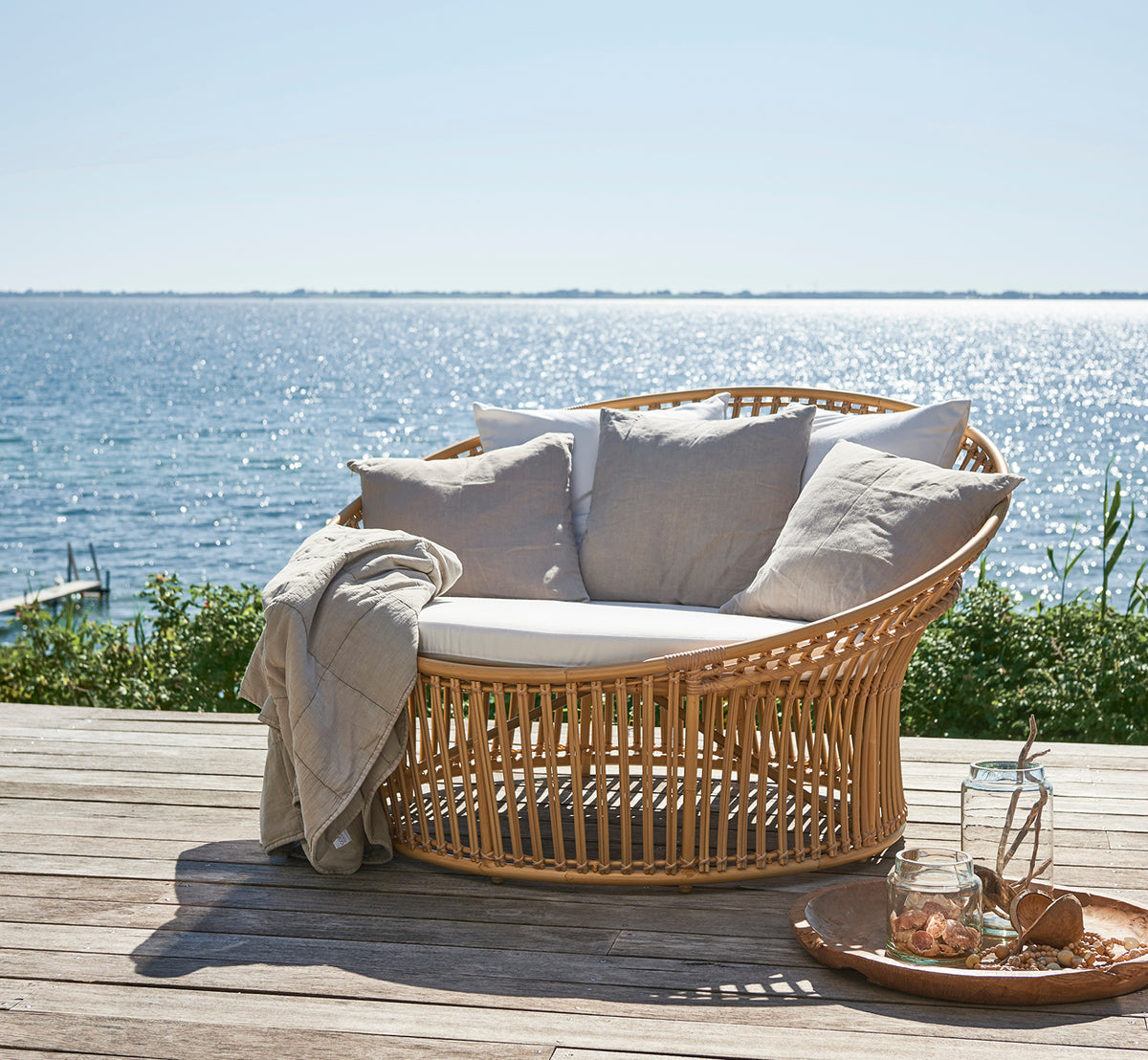 This is a lifestyle image of Olympia Nest Exterior in natural aluminium artfibre. On a beach pavilion with cushions and sea view.