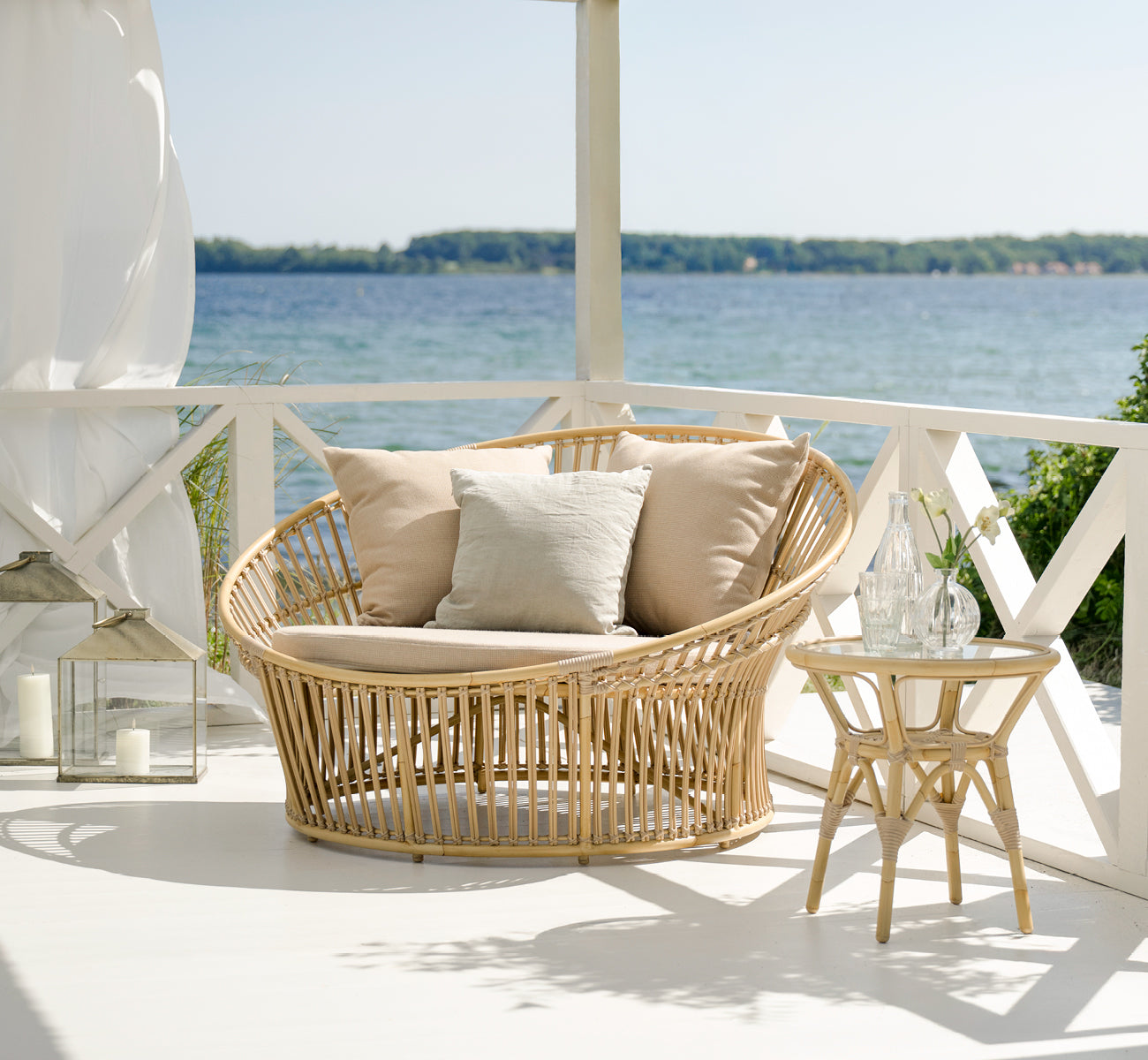 This is a lifestyle image of Olympia Nest Exterior in natural aluminium artfibre. On a seaside pavilion with linen cushions.