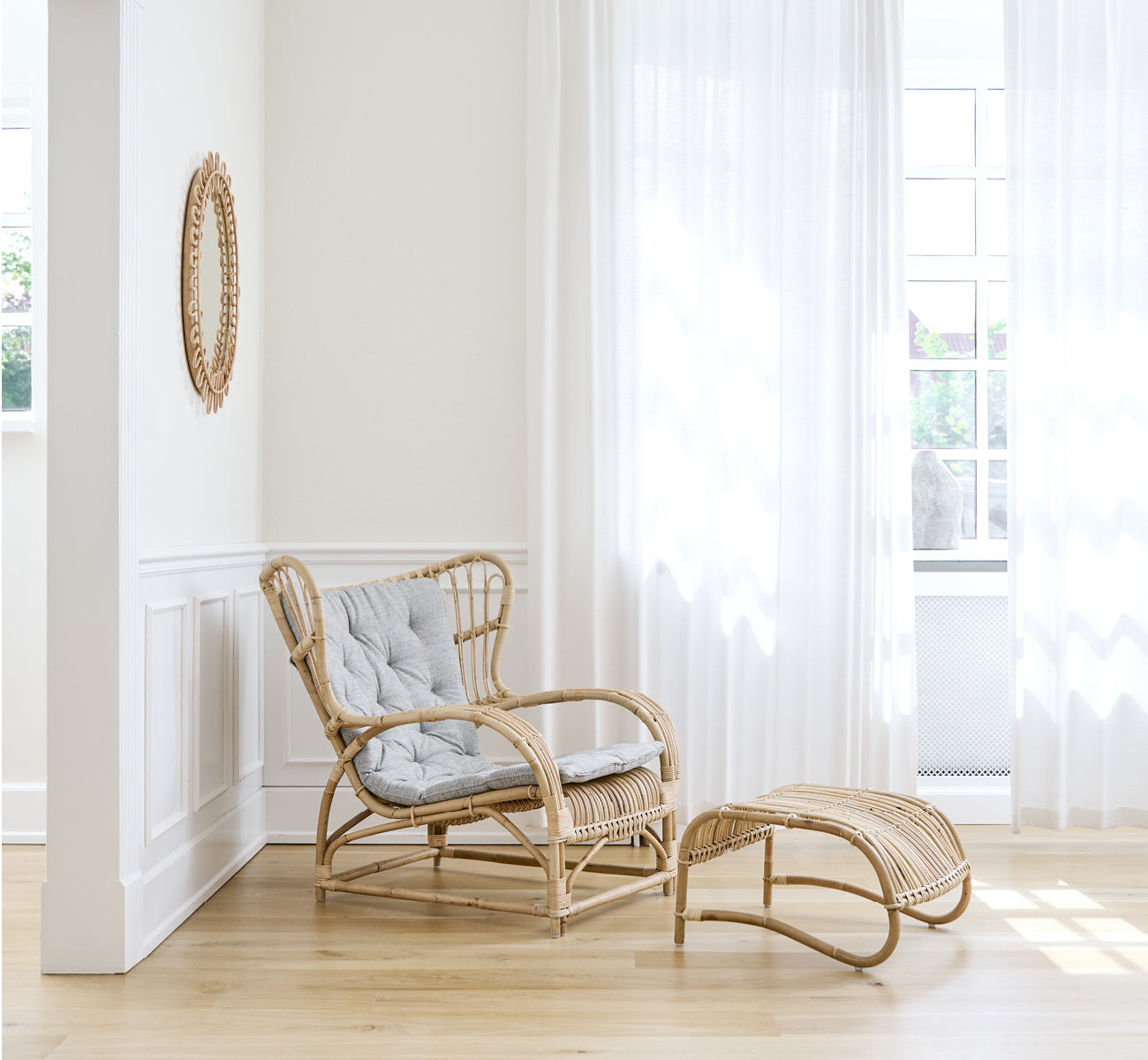 This is a lifestyle image of Teddy Lounge Chair in polished natural rattan. Placed on a herringbone floor by a curtained window with small mirror.