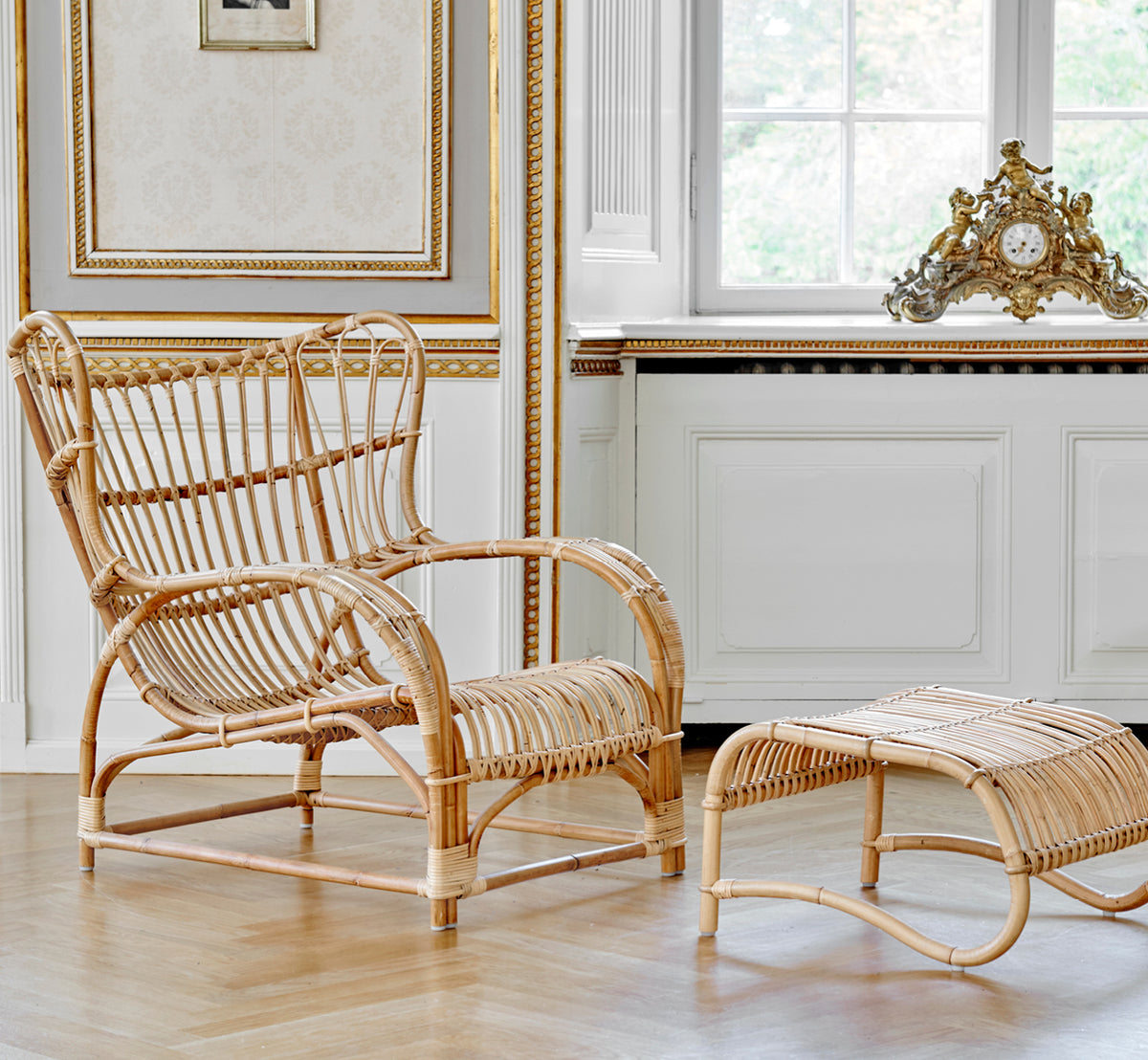 This is a lifestyle image of Teddy Lounge Chair in polished natural rattan. Placed on a herringbone floor by a curtained window with small mirror.