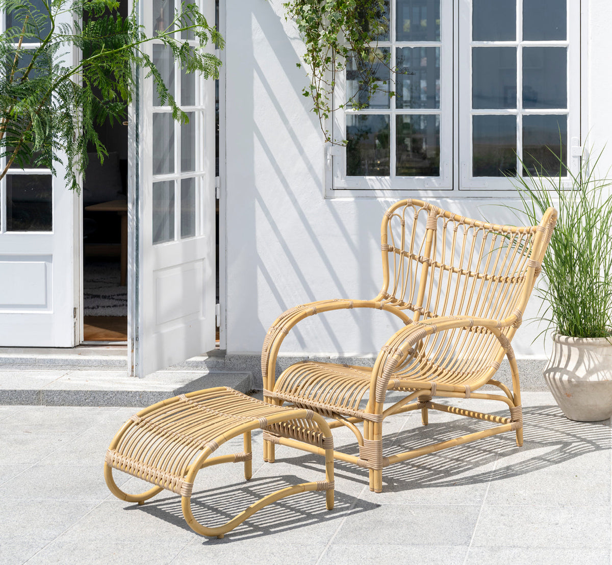 This is a lifestyle image of Teddy Exterior Lounge Chair in natural aluminium artfibre. Placed on a sunny paved terrace by a white-framed doorway.