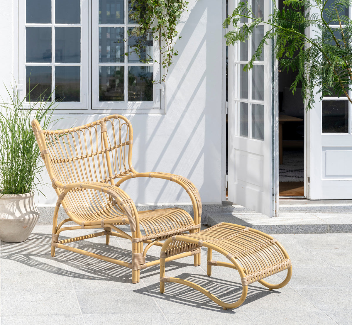 This is a lifestyle image of Teddy Exterior Lounge Chair in natural aluminium artfibre. Placed on a sunny paved terrace by a white-framed doorway.