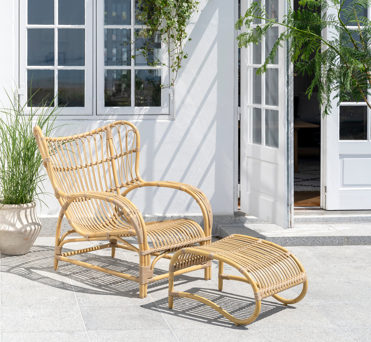 This is a lifestyle image of Teddy Exterior Lounge Chair in natural aluminium artfibre. Placed on a sunny paved terrace by a white-framed doorway.