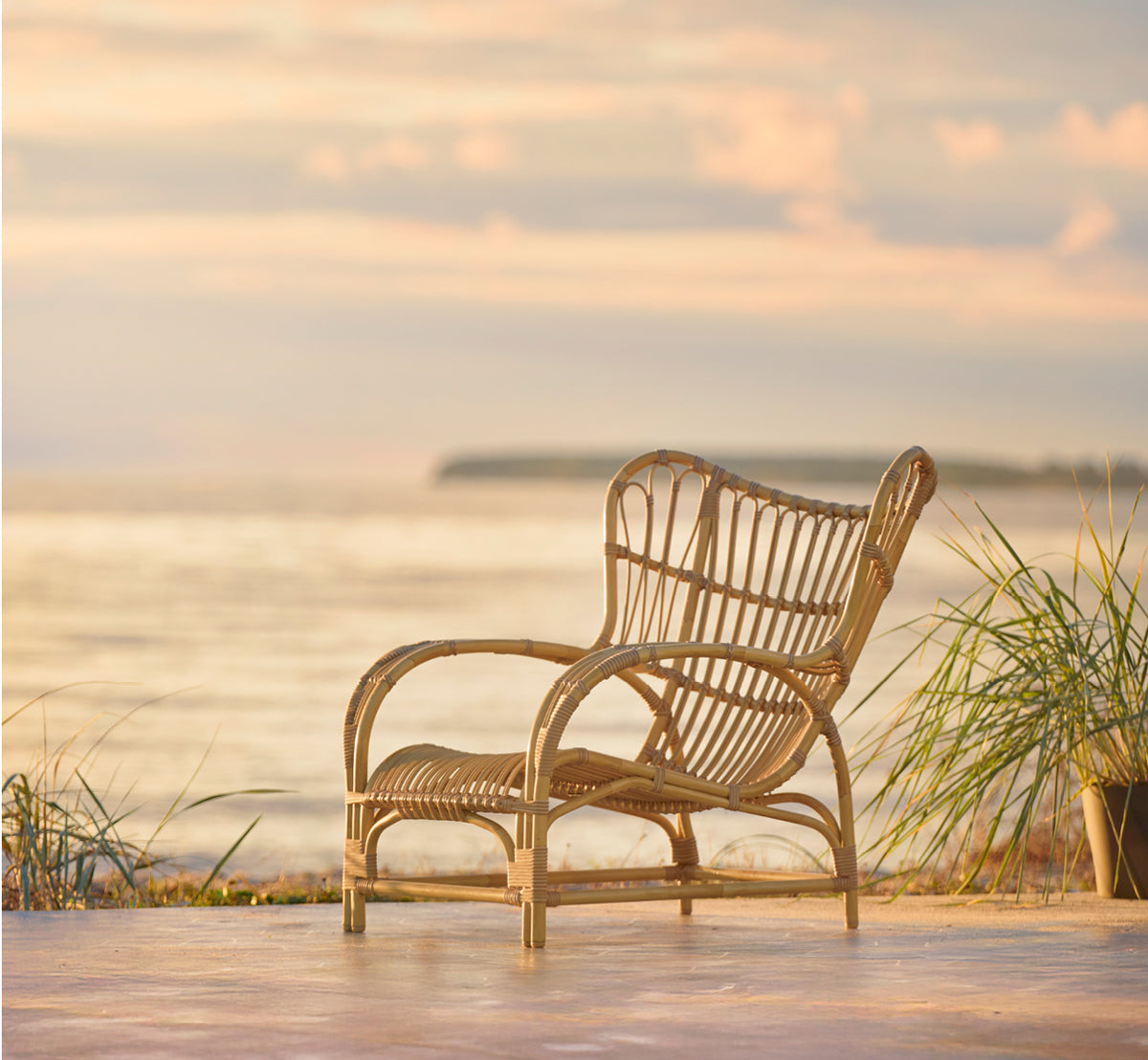 This is a lifestyle image of Teddy Exterior Lounge Chair in natural aluminium artfibre. Placed on a sunny paved terrace by a white-framed doorway.
