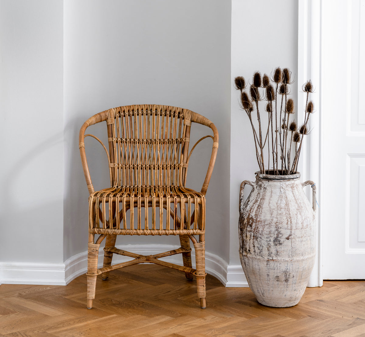 This is a lifestyle image of Robert Dining Chair in polished antique rattan. Placed beside a tall terracotta urn on a herringbone parquet floor.