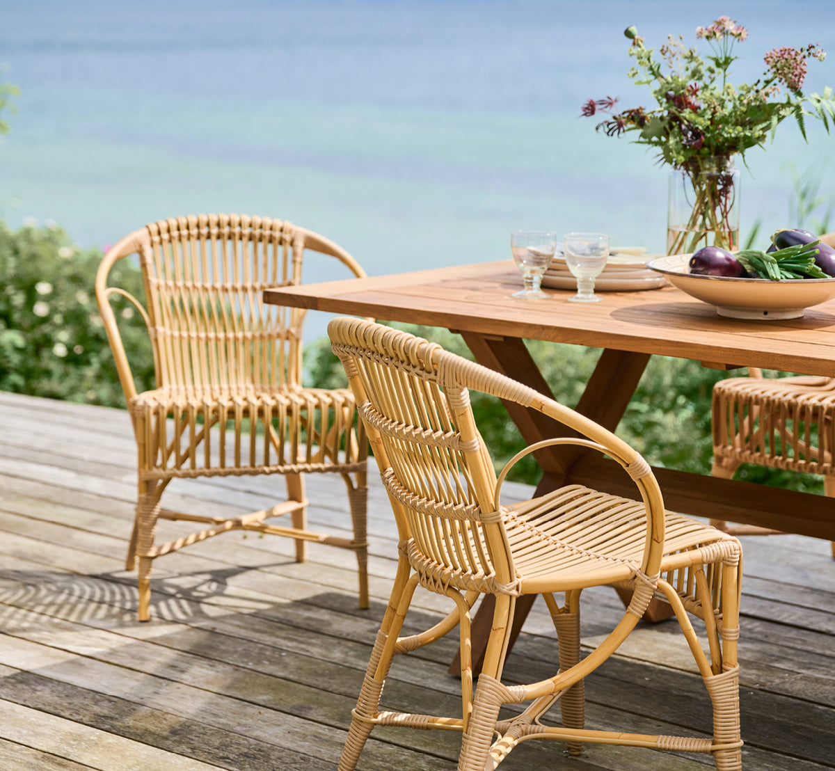This is a lifestyle image of Robert Exterior Dining Chair in natural aluminium artfibre. Set around a teak table on a wooden deck overlooking the sea.