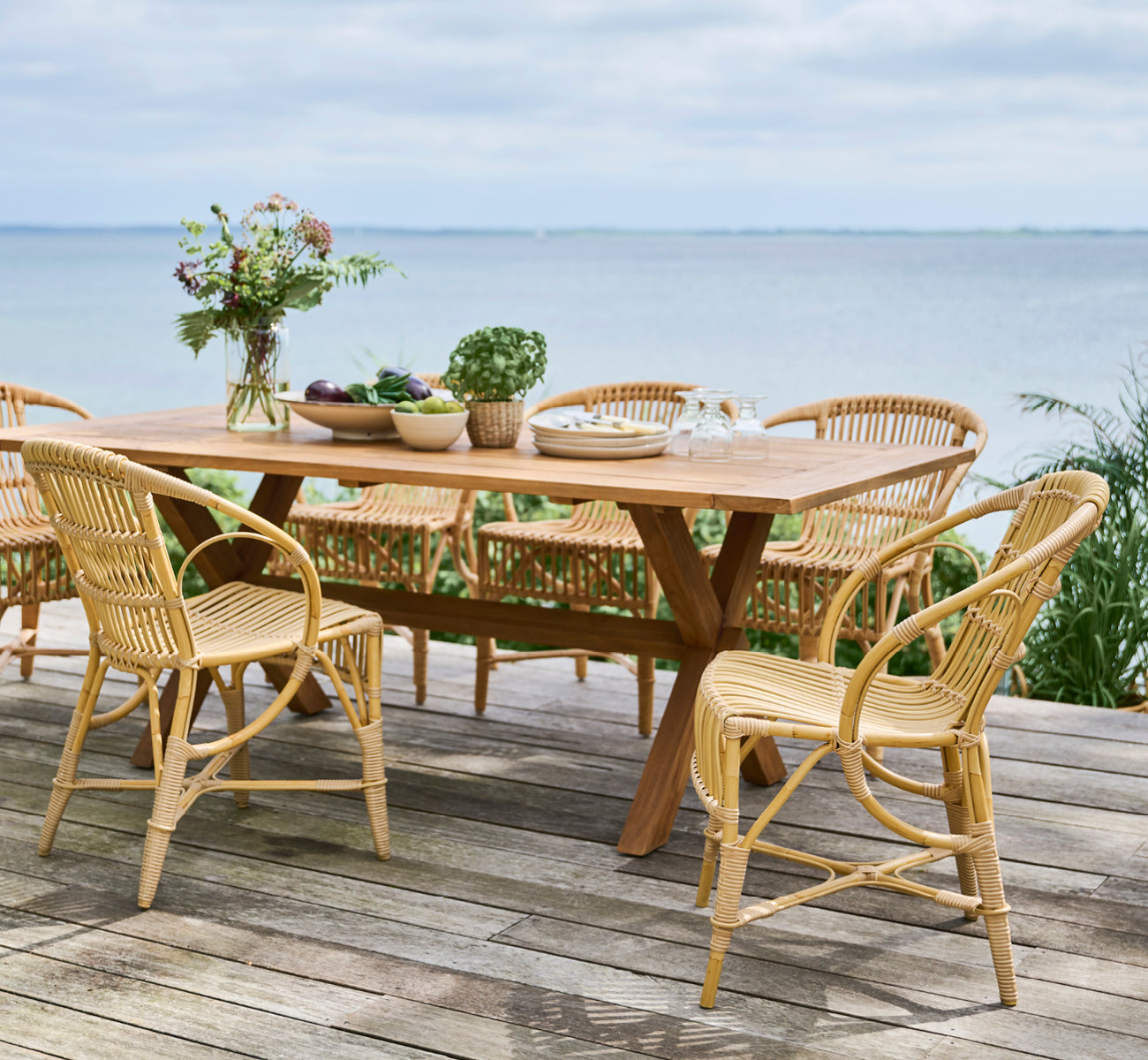 This is a lifestyle image of Robert Exterior Dining Chair in natural aluminium artfibre. Set around a teak table on a wooden deck overlooking the sea.