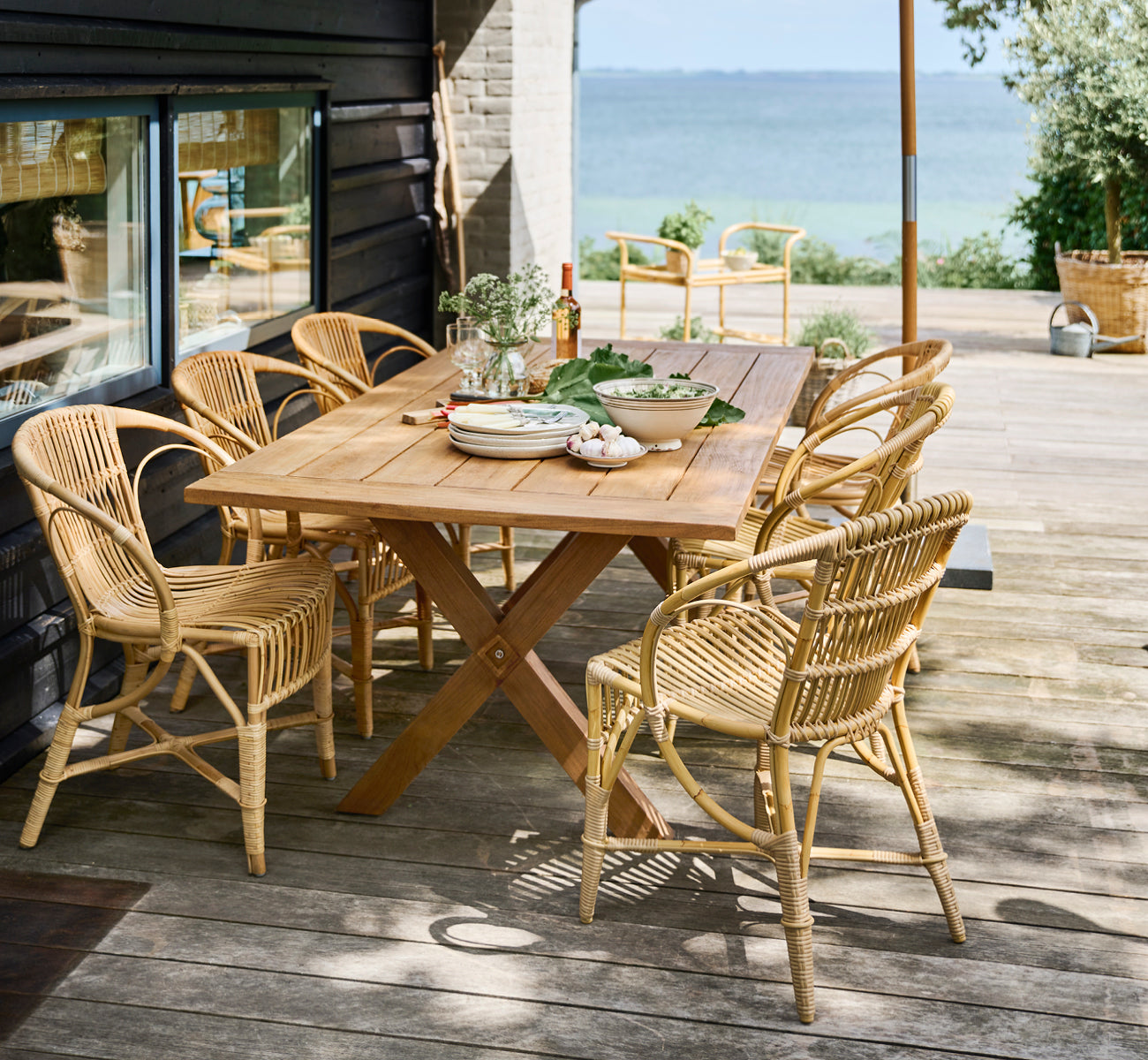 This is a lifestyle image of Robert Exterior Dining Chair in natural aluminium artfibre. Set around a teak table on a wooden deck overlooking the sea.
