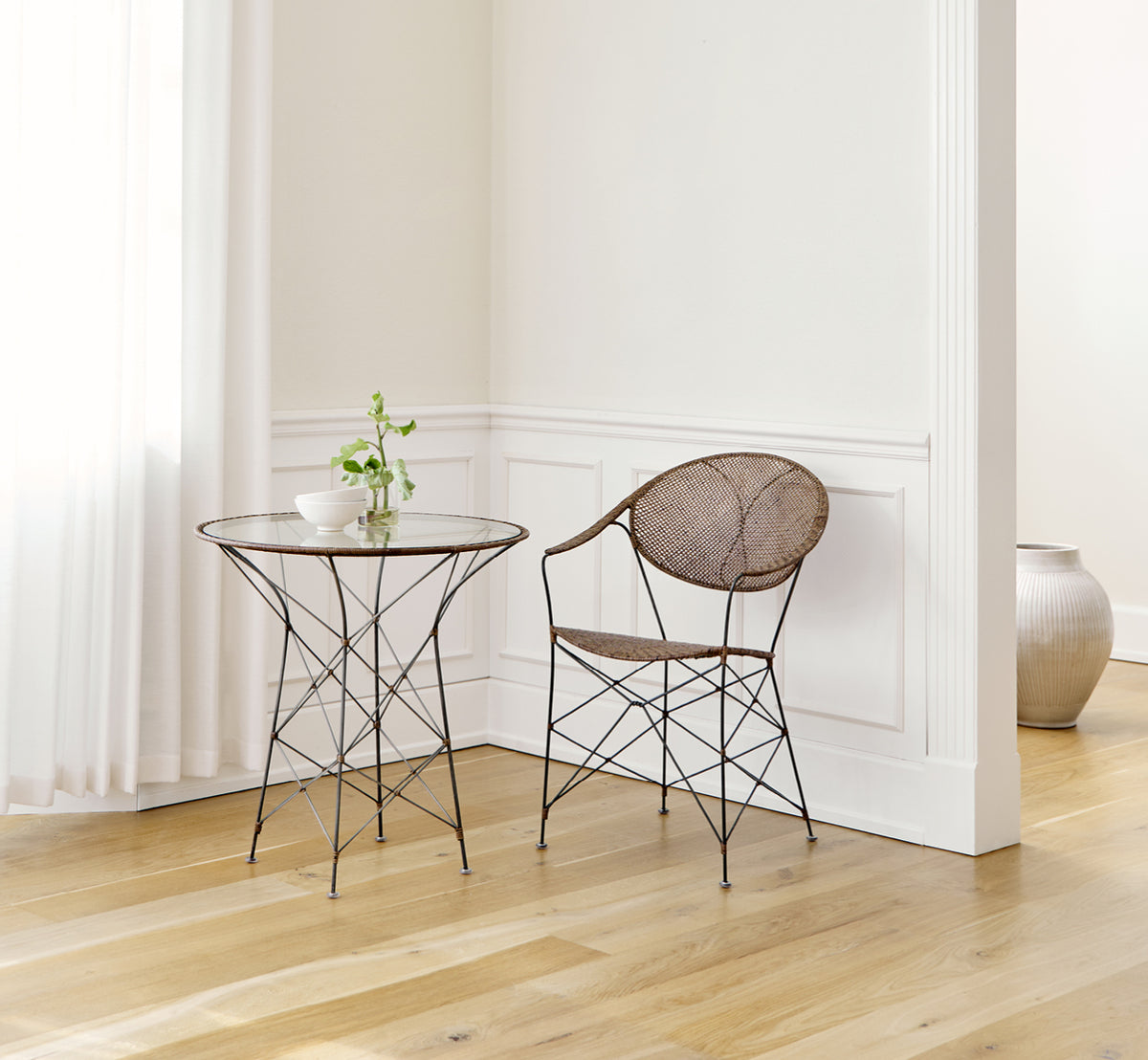 This is a lifestyle image of Funky Dining Chair in hazelnut rattan iron frame, in a bright interior with white panelled walls and herringbone parquet.