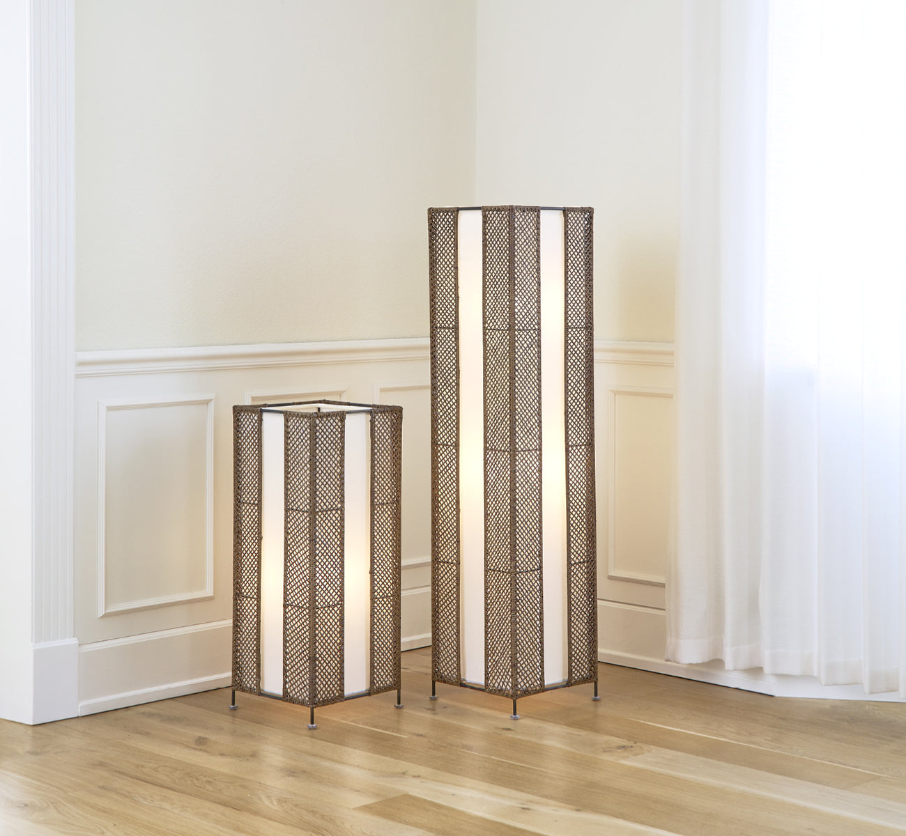 This is a lifestyle image of Yamakawa Lampshade | 125 cm in hazelnut rattan iron frame. Shown as two tall floor lamps in a bright panelled interior.